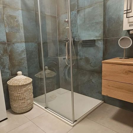 Dusche mit Glasabtrennung in einem Badezimmer mit grauen Fliesen, daneben ein Holzwaschtisch und ein Wäschekorb