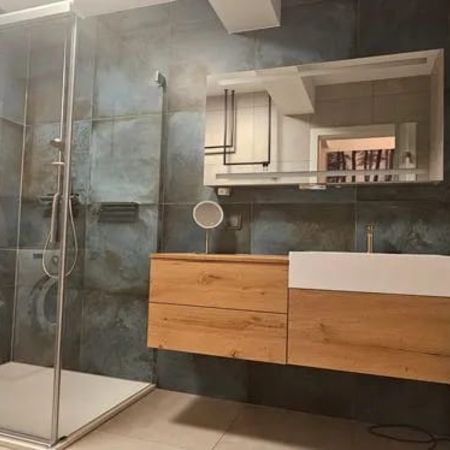 Dusche mit Glasabtrennung in einem Badezimmer mit grauen Fliesen, daneben ein Holzwaschtisch und ein Wäschekorb