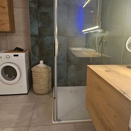 Dusche mit Glasabtrennung in einem Badezimmer mit grauen Fliesen, daneben ein Holzwaschtisch und ein Wäschekorb