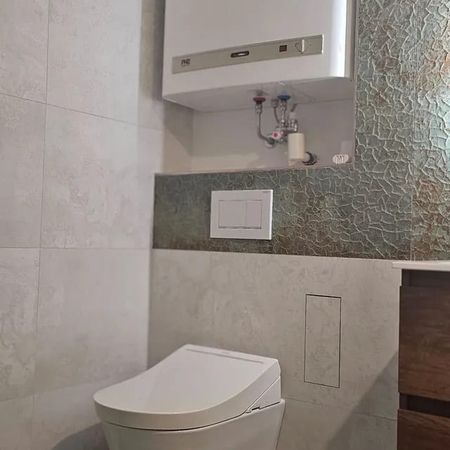 Moderne Wandtoilette mit verdecktem Spülkasten und weißem Boiler darüber in einem Badezimmer mit grauen Fliesen