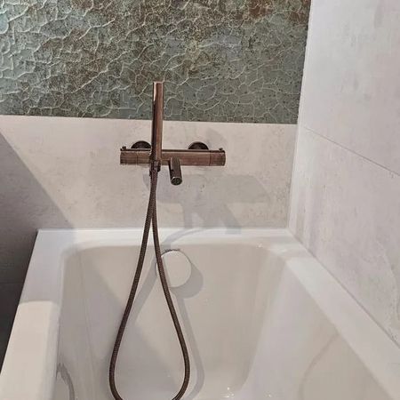 Weiße Badewanne mit brauner Wandarmatur und Handbrause vor einer Wand mit strukturierter grauer Fliesenfläche