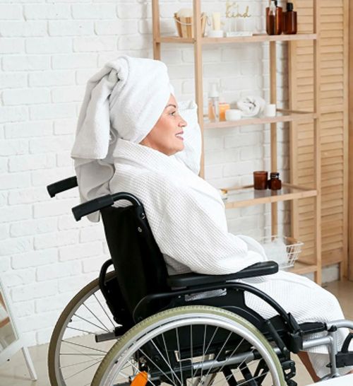 Person im weißen Bademantel und Handtuch auf dem Kopf sitzt im Rollstuhl in einem Badezimmer mit Holzregal und Pflegeprodukten