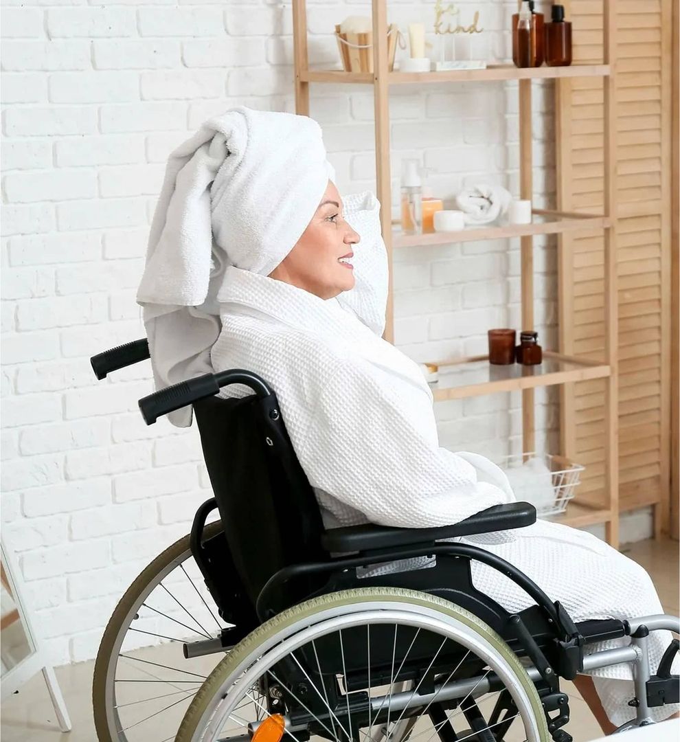 Person im weißen Bademantel und Handtuch auf dem Kopf sitzt im Rollstuhl in einem Badezimmer mit Regalen und Pflegeprodukten