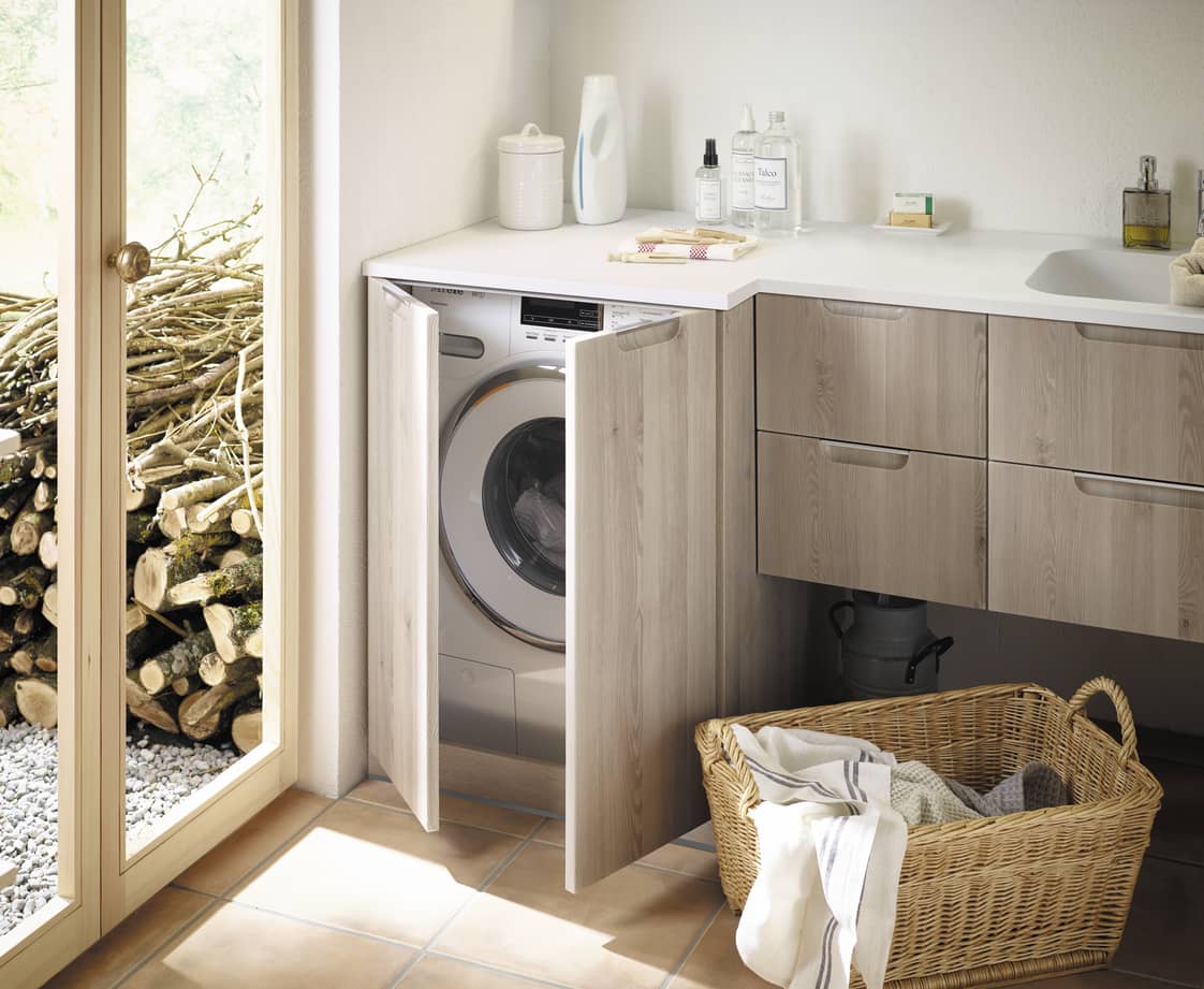 Waschmaschine in einer offenen Holzschranktür neben einem Waschbecken mit Seifen und Waschmitteln, Wäschekorb mit Kleidung auf dem Boden