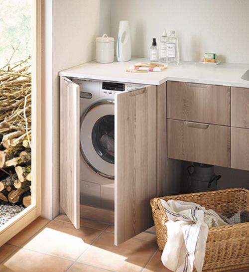 Waschmaschine in Holzschrank eingebaut, daneben Wäschekorb mit weißer Wäsche, helle Küchenzeile mit Waschmitteln