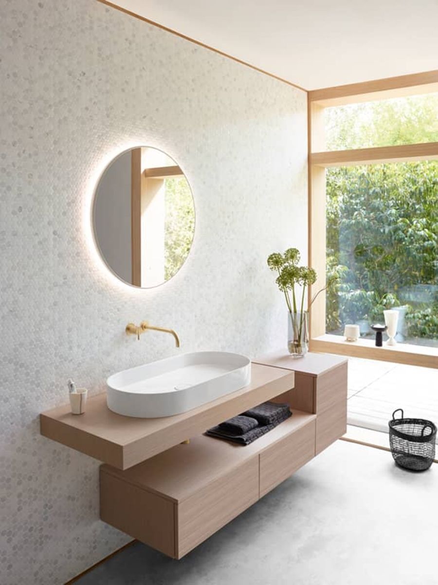 Modernes Badezimmer mit ovalem weißen Waschbecken auf hölzernem Waschtisch, rundem beleuchtetem Spiegel und großem Fenster mit Blick ins Grüne